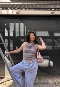 Top de halter marrón acanalado, pantalones de chándal anchos grises y un bolso rosa claro. El fondo presenta una pared oscura con sombras de una escalera.