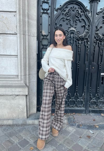 Jersey crema de corte amplio y hombros al descubierto; pantalones de pata ancha en cuadros marrones; botines de ante color marrón claro; de pie frente a puertas ornamentadas de color negro.