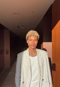 Νεαρός με σγουρά ξανθά μαλλιά, φορώντας ριγέ σακάκι και λευκό πουκάμισο, στέκεται σε διάδρομο με χαμηλό φωτισμό, όπου το ηλιακό φως ρίχνει σκιές πάνω του.