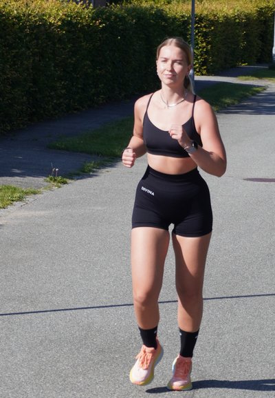 Sostén deportivo negro y pantalones cortos a juego, corriendo por un camino pavimentado. Lleva calcetas negras y zapatillas deportivas de color rosa claro. Ambiente soleado.