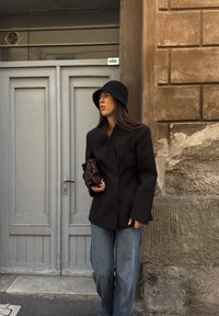 Čierny na mieru ušitý blejzer, široké modré džínsy, rebrovaná čierna látková čiapka, a tmavohnedá písacia taška. Hladká tkanina a štruktúrovaný strih.