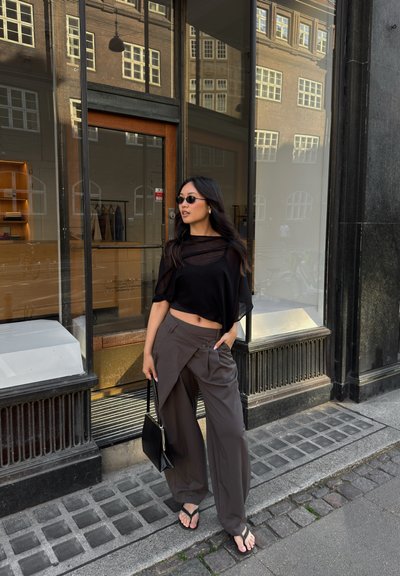 Top negro transparente, pantalones sueltos grises oscuros con diseño asimétrico, bolso negro y chanclas, contra el fondo de una vitrina.