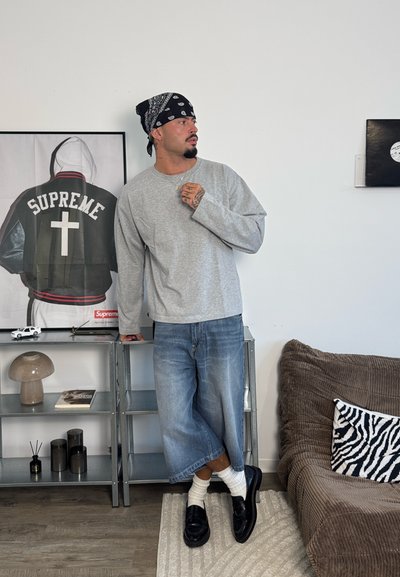 Hombre con bandana negra, camisa gris de manga larga y pantalones anchos de mezclilla, apoyado en una estantería metálica en un salón moderno.