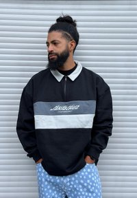 Homme avec barbe et chignon portant un sweat-shirt Santa Cruz noir et gris et un pantalon bleu à pois, debout devant un mur blanc rayé.