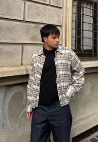 Camisa de botão com padrão xadrez cinza claro, mangas compridas e corte descontraído, usada sobre uma t-shirt preta. Calças de ganga escuras.