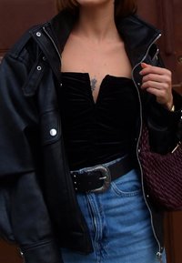 Veste en cuir noire avec un col montant, portée sur un haut noir froncé. Jupe en jean bleue, grosse boucle de ceinture, et sac à main tissé bordeaux.