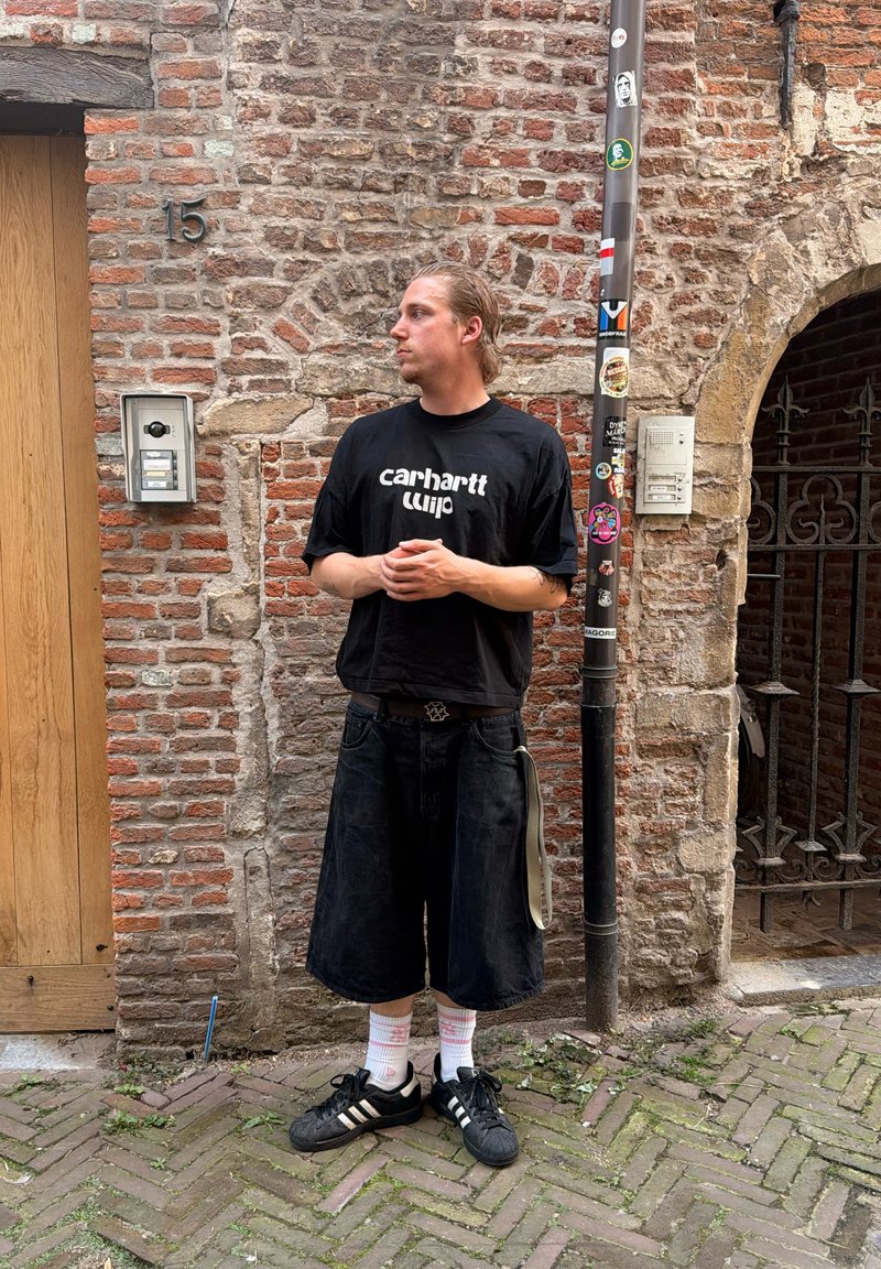 Jeune homme en t-shirt noir Carhartt et short ample, appuyé contre un mur en briques près d'une porte et d'un poteau avec des autocollants.