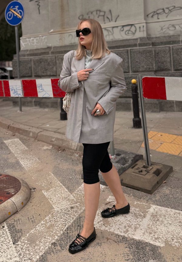 Femme portant un blazer gris oversize, des leggings noirs et des chaussures noires, debout dans une rue près de barricades rouges et blanches avec des graffitis sur le mur.