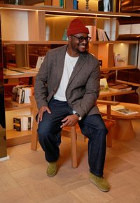 Veste en tweed marron sur un t-shirt blanc, jean bleu foncé et bottines en suède vert, assis sur une chaise en bois dans un intérieur moderne.
