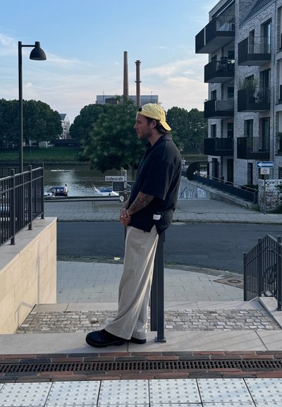 Camisa negra de manga corta, pantalones anchos beige, zapatos negros y gorra amarilla. De pie en unas escaleras cerca de un río y edificios modernos.