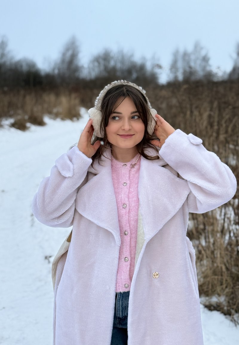 Jeune femme en pull rose et manteau blanc ajustant des cache-oreilles duveteux sur un chemin enneigé avec de l'herbe sèche et des arbres dénudés en arrière-plan.