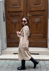 Beige trenchcoat med knytebelte i midjen, svarte ankelstøvler og ovale solbriller, som går foran utsmykkede tredører på en brosteinsgate.