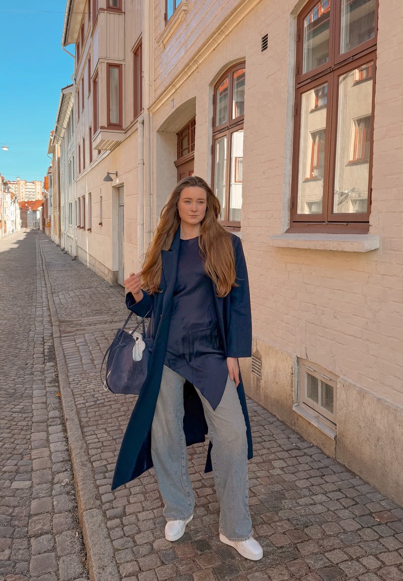 Femme aux longs cheveux portant un manteau bleu marine, un haut sombre et un jean bleu clair, debout dans une rue pavée, tenant un sac à main bleu marine avec un masque blanc attaché.