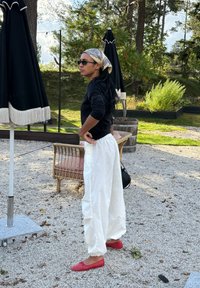 Γυναίκα που φοράει γυαλιά ηλίου, ένα μαντήλι με σχέδιο στο κεφάλι, μαύρο πουλόβερ, λευκό παντελόνι και κόκ κινα παπούτσια στέκεται έξω σε χαλίκι κοντά σε ψάθινα έπιπλα και κλειστές ομπρέλες.