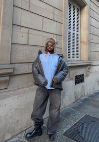 Veste grise gonflée sur une chemise bleu clair, associée à un jean foncé ample et des bottines noires. Accessoires comprenant un chapeau en fourrure et des lunettes de soleil.