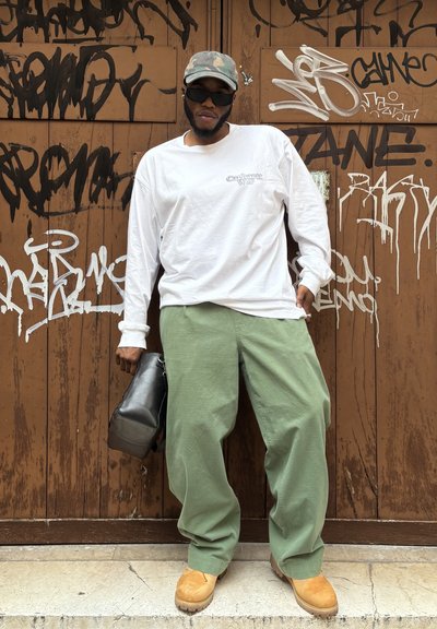 Camisa blanca de manga larga con logo, pantalones holgados verde claro, zuecos marrones y gorra gris camuflada. Sosteniendo un bolso negro contra un fondo de madera.
