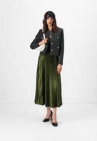 Chaqueta corta de cuero negra con botones metálicos, combinada con una falda de satén verde oliva con plisados. La modelo sostiene un bolso blanco de forma curvada y lleva tacones negros.