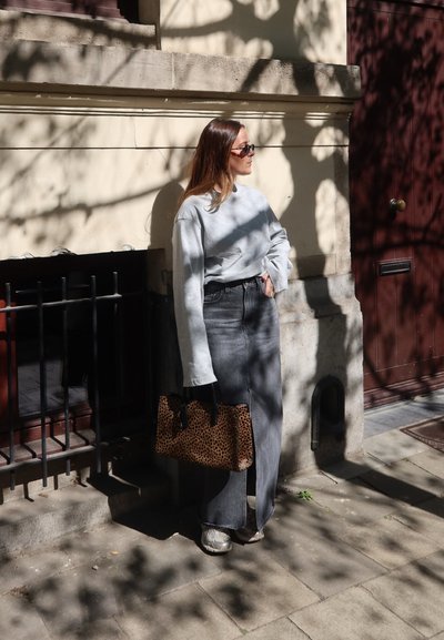 Sudadera gris, vaqueros negros de talle alto, bolso con estampado de animal. La luz del sol y las sombras crean un patrón en la pared detrás.