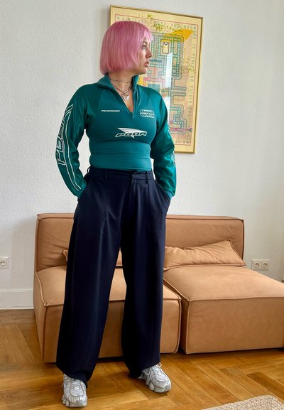 Camiseta de manga larga azul verdoso con cremallera y logotipos, combinada con pantalones sueltos negros y zapatillas grises, sobre un fondo neutro.