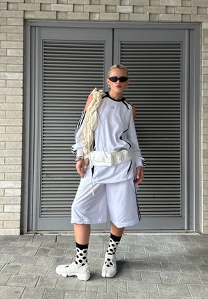 White athletic outfit featuring black accents, comprising a long-sleeve top and knee-length shorts. Accessories include a bum bag and polka dot socks.
