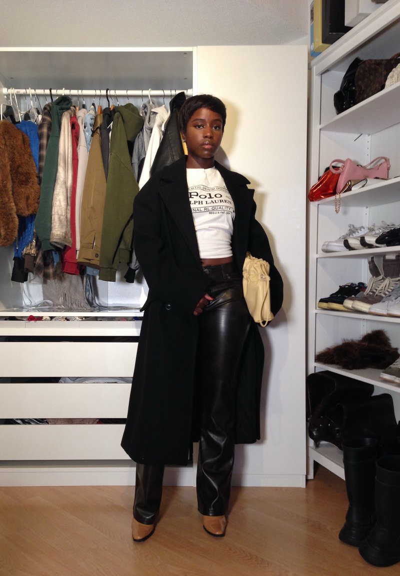 Manteau noir, T-shirt graphique blanc, pantalon en cuir noir, sac beige ; sol en bois en arrière-plan, placard organisé avec divers vêtements et chaussures.