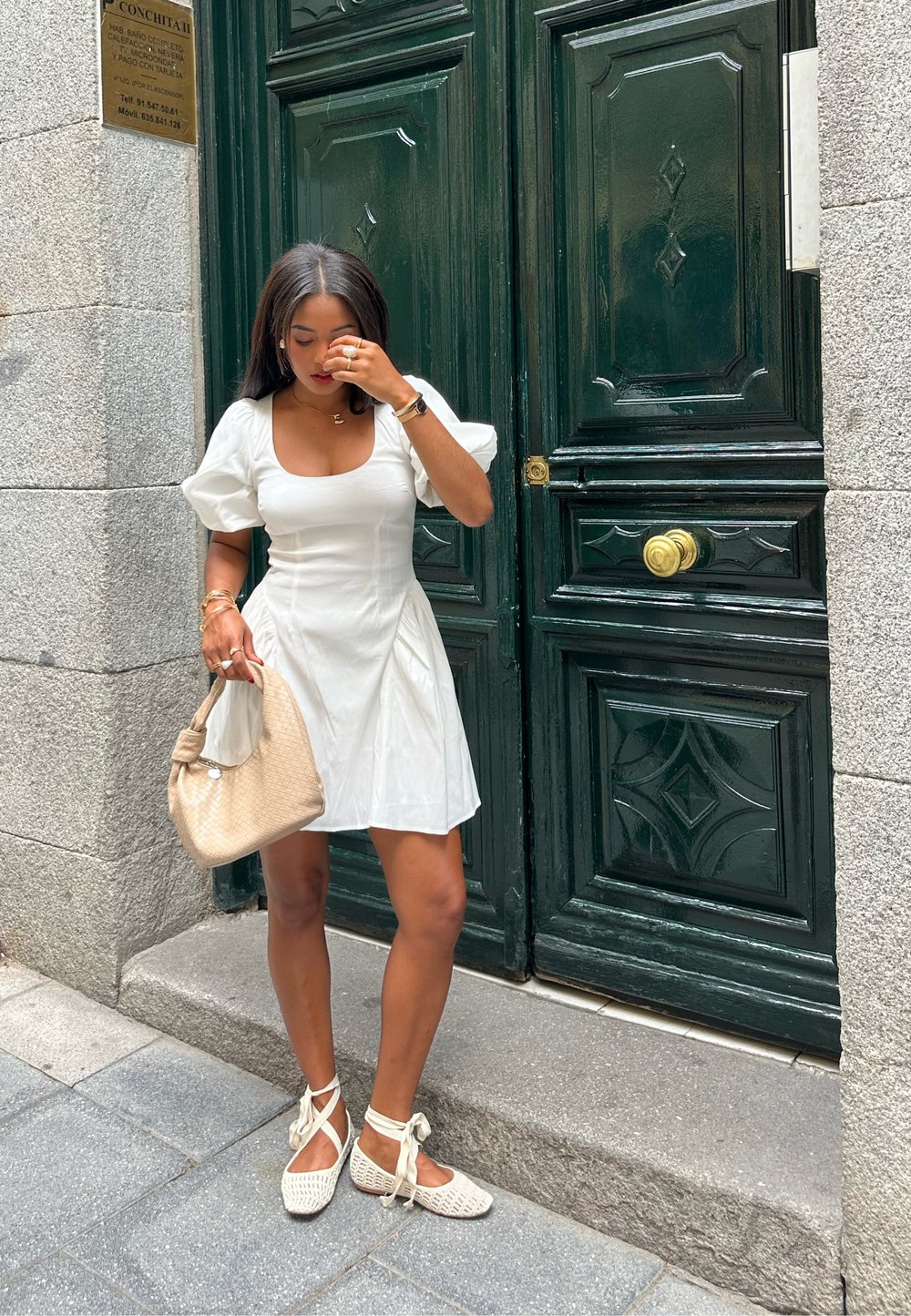 Vestido curto branco com mangas bufantes, combinado com uma Bolsa de palha bege e espadrilhas de atacadores de cor clara, em frente a portas verdes.