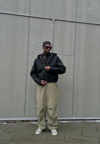 Felpa nera con zip e vestibilità comoda, abbinata a pantaloni cargo beige larghi e sneaker bianche. Lo sfondo presenta pannelli grigi.