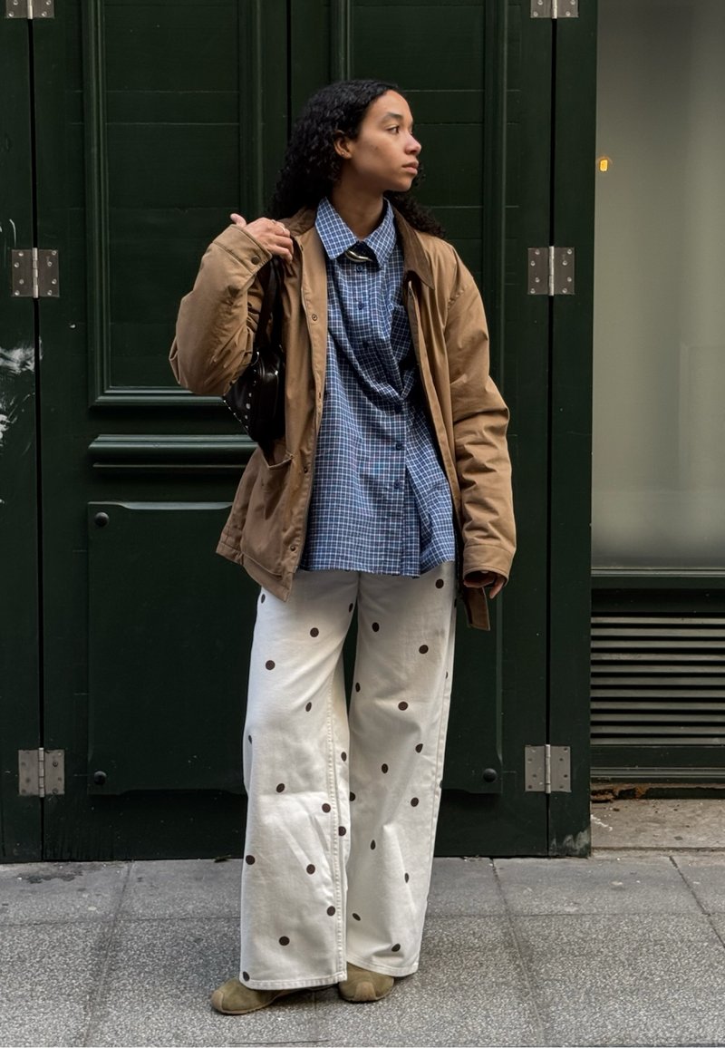Jeune femme debout sur un trottoir en ville, portant une veste marron, une chemise à carreaux bleus, un pantalon blanc à pois marron, tenant un sac noir.