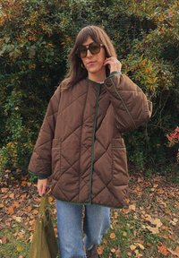 Barna steppelt dzseki zöld szegéllyel, két elülső zsebbel és laza fazonnal, világoskék öltözet felett viselve, zöld táskával párosítva.