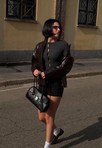 Crna kožna torba, sivi top s detaljima gumba, smeđa sjajna jakna, crne kratke hlače, bijele čarape i crne cipele s studovima. Urbano okruženje.