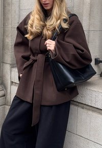 Vêtement d'extérieur marron ceinturé avec manches larges, associé à un sac à main en cuir noir. Pantalon noir ample en tissu texturé, fond neutre.