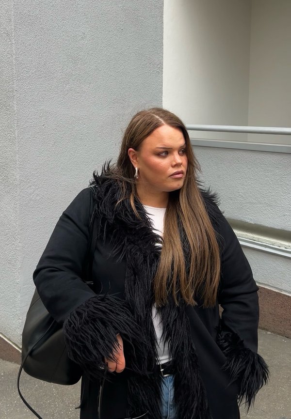 Młoda kobieta z długimi brązowymi włosami, ubrana w czarny płaszcz z futrzanym wykończeniem, biały top i jeansy, stojąca na zewnątrz przy ścianie budynku.