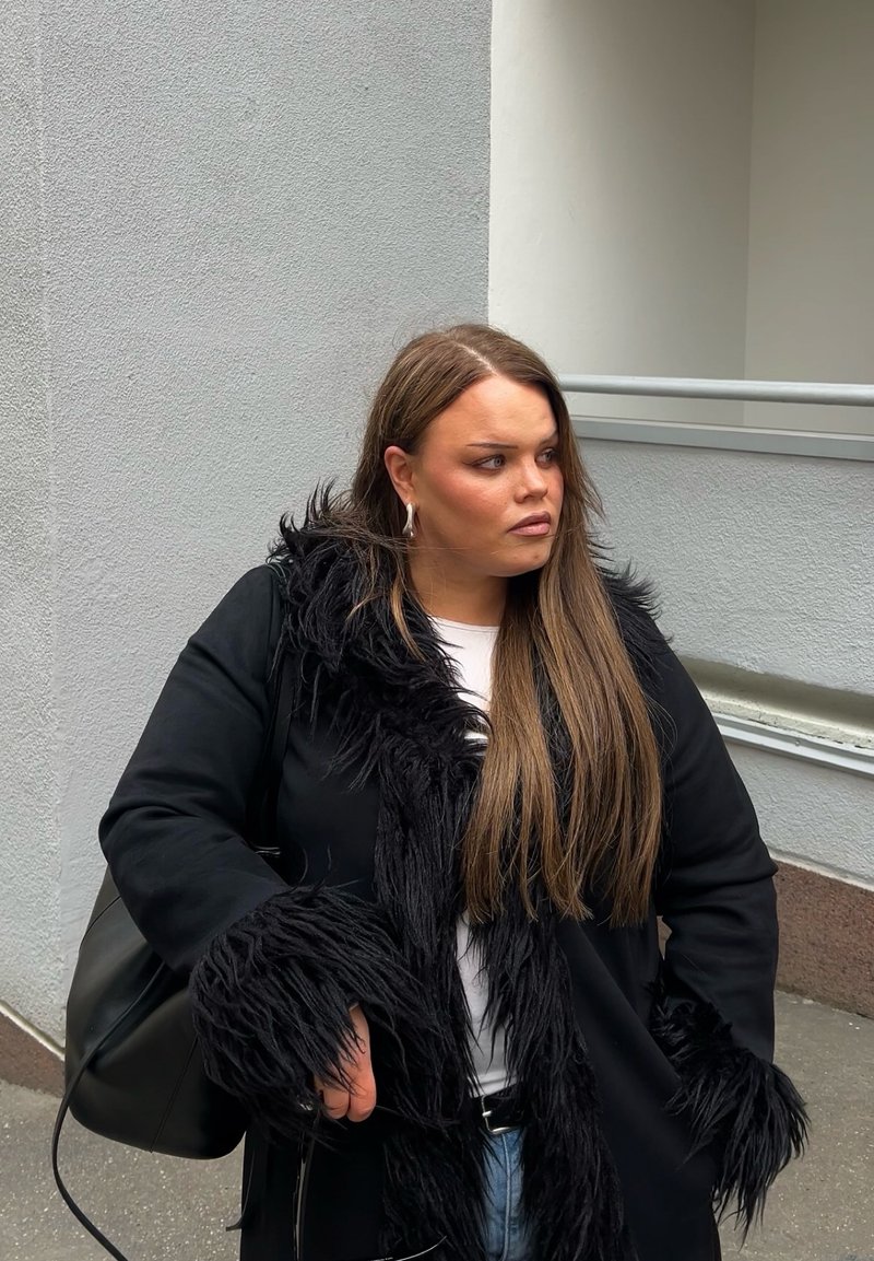 Junge Frau mit langen braunen Haaren, die einen schwarzen Mantel mit Pelzbesatz, ein weißes Oberteil und Jeans trägt, steht draußen nahe einer Gebäudeaußenwand.