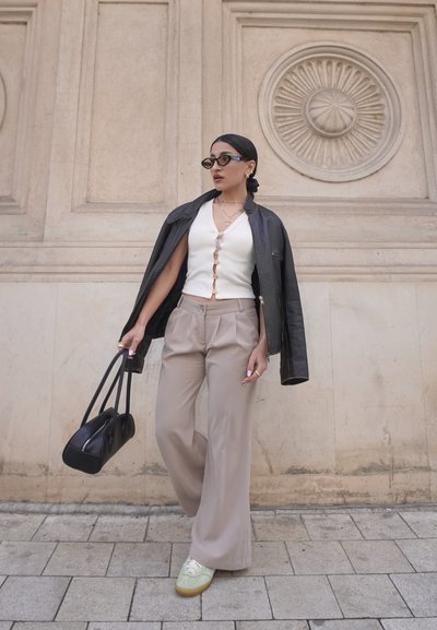 Chaqueta de cuero negra sobre una blusa blanca de canalé, pantalones beiges de pierna ancha, zapatillas verdes y beiges, bolso negro, con un fondo de pared intrincado.