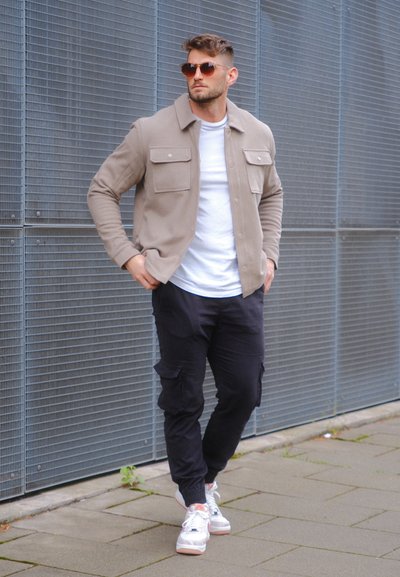 Chaqueta abotonada color beige claro con dos bolsillos frontales, llevada sobre una camisa blanca y unos pantalones cargo negros, combinada con zapatillas blancas y gafas de sol.