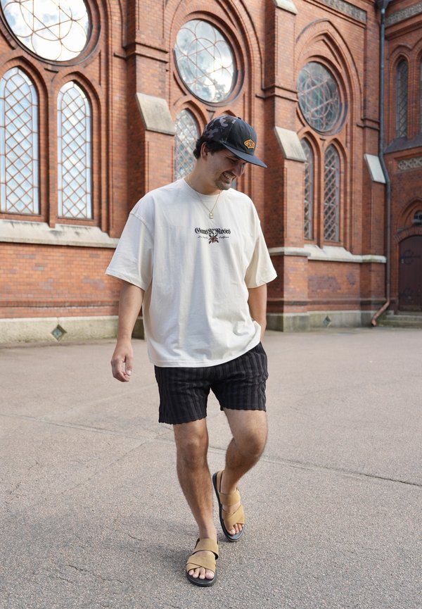 Homme portant une casquette noire, un t-shirt blanc Guns N' Roses, un short noir et des sandales marchant sur un trottoir à côté d'un bâtiment en briques rouges avec des fenêtres cintrées.