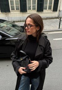 Čierny trenčkot cez čierny rolák, doplnený džínsami. Drží malú čiernu kabelku so zlatými doplnkami. Na tvári má okuliare.