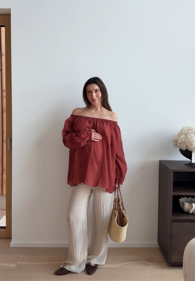 Femme debout à l'intérieur portant un chemisier rouille à épaules dénudées, un pantalon large crème, tenant un sac à main tissé et touchant doucement son ventre.