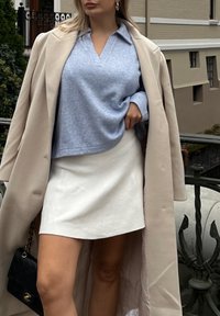 Ανοιχτό μπλε, υφασμένο πουλόβερ σε σχήμα V κάτω από ένα μπεζ παλτό, συνδυασμένο με μια λευκή μίνι φούστα. Μικρή μαύρη τσάντα με λεπτομέρεια αλυσίδας.