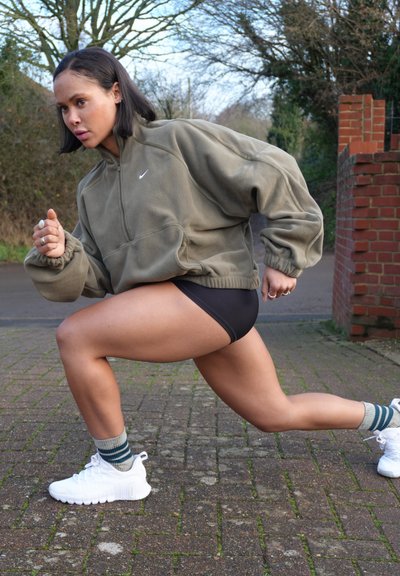 Joven estirando las piernas en una zancada al aire libre sobre un camino de ladrillos, vistiendo una chaqueta verde con cremallera, pantalones cortos negros, calcetas a rayas y zapatillas blancas.
