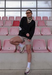 Chemise noire à manches longues avec détail graphique, short beige, écharpe à motifs, lunettes de soleil et baskets bicolores sur un siège de stade rose.