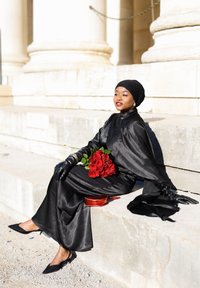 Γυναίκα με μαύρο φόρεμα, μαντήλι στο κεφάλι και γάντια, καθισμένη σε λίθινα σκαλοπάτια, κρατώντας κόκκινα τριαντάφυλλα με κόκκινη τσάντα δίπλα της κάτω από έντονο ηλιακό φως.