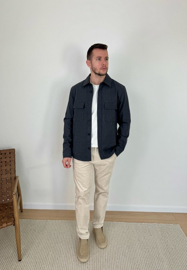 Uomo con una camicia scura abbottonata sopra una t-shirt bianca, pantaloni beige e scarpe beige, in piedi su un tappeto accanto a una sedia intrecciata.