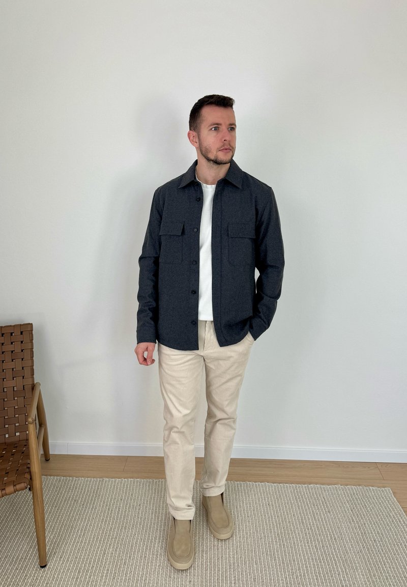 Homme portant une chemise sombre à boutons par-dessus un t-shirt blanc, un pantalon beige et des chaussures beiges, debout sur un tapis à côté d'une chaise en osier.
