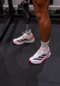 White trainers with black stripes and red accents, worn with white socks. Partial view of a prosthetic limb next to the trainers.