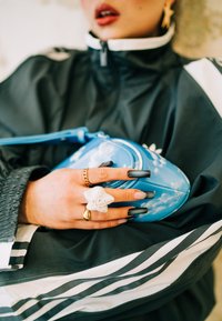 Bolsa de crossbody azul com padrão de nuvens, textura brilhante, fecho zip, segurada por uma mão adornada com anéis e unhas longas e escuras.