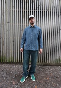 Chemise en denim à rayures bleues, jean bleu ample et baskets vertes avec des accents blancs. Le mannequin se tient devant un fond en lattes de bois.