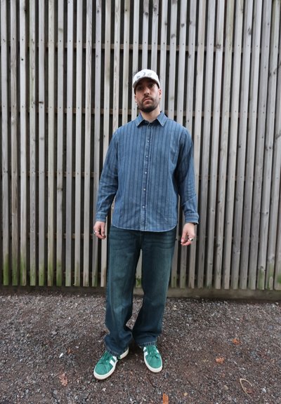 Camisa de mezclilla con rayas azules, jeans azules holgados y zapatillas verdes con detalles blancos. La modelo está de pie frente a un fondo de listones de madera.