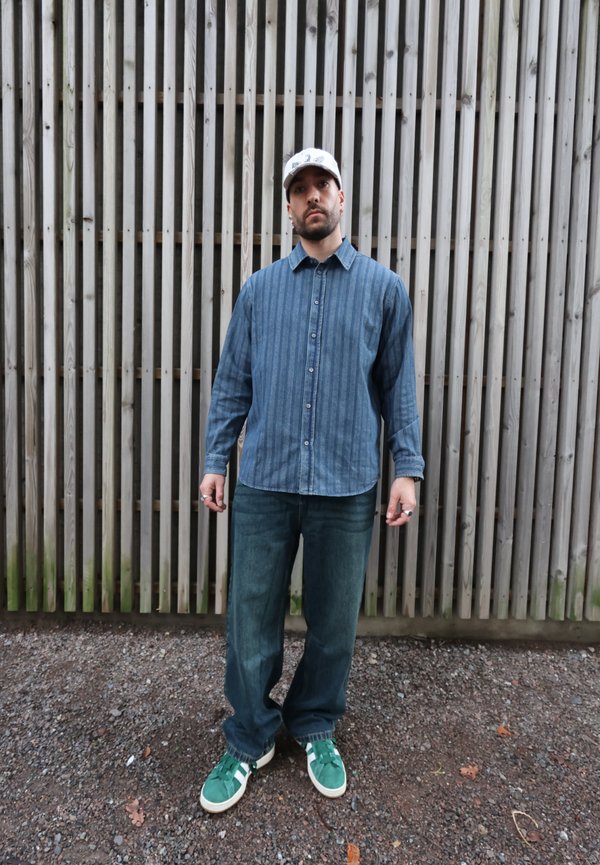 Camicia di jeans a righe blu, jeans blu larghi e sneakers verdi con dettagli bianchi. Il modello è in piedi davanti a uno sfondo di doghe di legno.