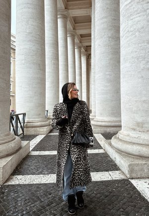 Mujer con un abrigo de estampado de leopardo y capucha negra caminando entre altas columnas de mármol blanco sobre un pavimento de piedra con patrones.